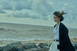 © SHOTPRIME STUDIO - Woman on a rocky shoreline, wind in her hair, wearing a white shirt and dark jacket, gazing toward the sea beneath moody clouds, a fashion mood by the coast