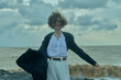 © SHOTPRIME STUDIO - Female professional in a blazer and white shirt on a windy beach, holding her coat open as waves crash, capturing bold fashion and coastal movement against a vast ocean backdrop