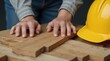 © Alex Pios - Construction worker's hands resting on wooden boards with a yellow hard hat in the foreground, highlighting a safe work environment.