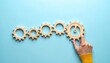 © Qaisar  - Six interlocking wooden gears in diagonal line on light blue surface, hand touching largest gear on right.