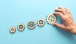 © Qaisar  - Six interlocking wooden gears in diagonal line on light blue surface, hand touching largest gear on right.