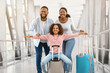 © Prostock-studio - Happy Loving Family In Airport. Portrait of joyful excited black girl sitting on suitcase and laughing, spreading arms imitating plane, smiling cheerful parents posing, standing in terminal hall