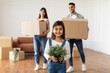 © Prostock-studio - House Move And Relocation Concept. Portrait Of Cheerful Family Of Three Housing, Selective Focus On Happy Smiling Little Girl Holding Plant, Parents Carrying Cardboard Boxes In Blurred Background