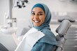 © Prostock-studio - Stomatologic Treatment. Portrait Of Happy Young Islamic Woman In Hijab Sitting In Dentists Chair In Clinic, Muslim Female Patient Waiting For Check Up With Orthodont, Looking At Camera