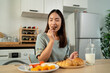 © Kawee - Asian young woman eating healthy fruits and bakery in kitchen at home.