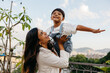 © (JLco) Julia Amaral - Mother lifts happy child outdoors with a city view in the background