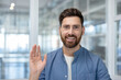 © Liubomir - Friendly bearded man looking at camera, smiling and waving hand, making a virtual greeting during a video call or online meeting from a modern office environment