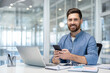 © Liubomir - Entrepreneurial man sitting at modern desk in office working on a laptop, holding a mobile phone, and smiling while managing communications and productivity