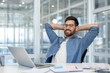 © Liubomir - Young man wearing eyeglasses feeling relaxed and smiling, leaning back in his chair with hands behind head, taking a break from working with laptop at modern office