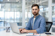© Liubomir - Bearded man sitting at a desk with a laptop, looking directly at the viewer in a modern office, representing concepts of remote work, business, technology, and focus