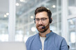 © Liubomir - Man wearing glasses and a headset, smiling while looking at a laptop, representing helpful communication and professional customer support in an office setting