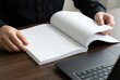 © Evgenia - Hands of a person holding an open blank notebook on a wooden desk beside a laptop, creating a workspace atmosphere for writing or brainstorming ideas