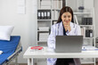 © Wasan - Young female doctor smiling while working on a laptop in medical office, with a stethoscope and medical records on the desk and a hospital bed nearby.