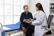 © Wasan - Asian female doctor wearing white coat and stethoscope consulting senior patient sitting on hospital bed, taking notes on clipboard in clinic room.
