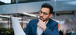 © Prostock-studio - A man wearing glasses looks thoughtfully at a document in a modern office. He is dressed in a suit and has a serious expression as he considers the information.