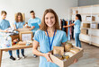 © Prostock-studio - Positive female volunteer in blue uniform holds a cardboard box filled with food donations. She smiles at the camera while working alongside diverse friends in a busy charity center.