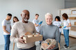 © Prostock-studio - A happy mixed race senior couple smiles at the camera while holding a donation box and string bag filled with food. In the background, volunteers are busy packing food for those in need.