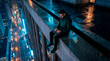 © Rendara Studio - Hacker man working on a laptop on a rainy skyscraper ledge with city lights below