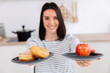 © Xavier Lorenzo - Young happy woman showing donuts and apple on plates, making healthy food choices in kitchen, promoting balanced nutrition and lifestyle