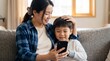 © Meow Creations - Cheerful asian mom showing content on cellphone to young son in living room