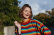 © SHOTPRIME STUDIO - Young woman in her early 20s wearing a colorful striped sweater smiling and laughing outdoors, holding yellow sunglasses; candid lifestyle portrait, autumn fashion casual outfit in park sunlight.