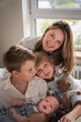 © Sofia - A mother embraces her three young sons, smiling warmly by the window as they sit together on the bed