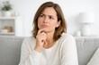 © hobonski - Thoughtful Woman Sitting On Sofa With Concerned Expression. A woman sits on a sofa touching her chin, showing uncertainty and inner tension while reflecting on a difficult decision in a calm home.