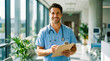 © NikProArt - Professional Male Healthcare Worker Smiling with Clipboard in Modern Hospital Corridor.