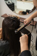 © vfhnb12 - Professional hairstylist using a flat iron to straighten long dark hair in a beauty salon.