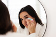 © Xavier Lorenzo - Happy woman wearing bathrobe using reusable cotton pad for removing makeup, enjoying morning skincare routine in front of bathroom mirror