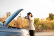 © Sirirat Makprasert - A woman showing frustration and stress by holding her head while looking at the open hood of her broken-down car on a quiet road during sunset.