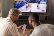 © ty - Two friends holding smartphone with sports bets app while watching basketball match at home in living room.