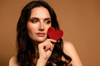 © Beauty Hero - Beautiful brunette woman holding red heart decoration on beige background with elegant makeup and stylish appearance