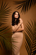 © Beauty Hero - Elegant young woman in a beige dress posing amidst palm leaves on a soft vertical background