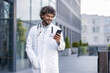 © Tetiana - Smiling young Indian male doctor standing in white coat on the street and using mobile phone