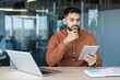© Liubomir - Young indian man in a modern office environment focused and thoughtfully reviewing information on a digital tablet, demonstrating concentration and problem-solving skills at work