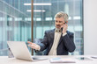 © Liubomir - Mature businessman experiencing stress during a frustrating phone call while looking at his laptop, dealing with problems in a modern office environment