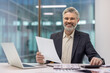 © Liubomir - Experienced businessman with gray hair and beard sitting at his office desk, holding a document, and using a calculator while looking at the camera with a confident smile