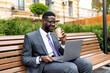 © Prostock-studio - Corporate african american executive in formal wear sitting on bench near office center with laptop and takeout coffee during lunch break. Happy black IT manager getting ready for business meeting