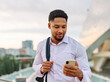 © Lumos sp - Portrait of a young businessman man using a smartphone mobile phone walking down the street, surrounded by moder corporate office buildings and modern city architecture