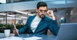 © Prostock-studio - A man in a suit sits at an office table focused on his laptop while holding a tablet. He appears to be analyzing data or documents. A cup and a notepad are on the table.