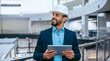 © Prostock-studio - A construction manager dressed in a suit and hard hat stands indoors in a factory. He is looking at a tablet and smiling. The background shows industrial equipment and bright windows.