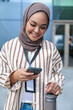© Jovo Jovanovic/Stocksy - Smiling woman in hijab checks phone, holding water bottle.