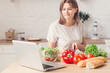 © zadorozhna - Woman cutting vegetables on salad and view recipes in laptop on kitchen.