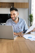 © fizkes - Smiling Hispanic man sit at table at home office using laptop working remotely, reviewing documents, analyzing business data, preparing reports, learn financial charts or graphs, doing online research