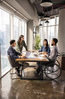 © fizkes - Diverse business team including young African professional in wheelchair discussing project marketing strategy at table, analyzing sales reports together, meeting for brainstorming. Vertical shot