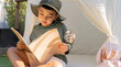 © Kunlathida - Cute Asian boy with hiking hat sitting in a tent teepee with book and magnifying glass