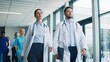 © VAKSMANV - Confident medical team walking through clinic hallway preparing for upcoming operation. Focused doctors carrying tablets and reviewing tasks while moving together. Professionals showing determination.