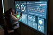 © artitwpd - Young Indian and senior Asian woman review interactive charts on a large digital screen in a modern office, showcasing corporate diversity, tech leadership, data analysis, and collaborative strategy.