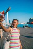 Carefree young Black woman laughing while taking selfie on pier, showing pure joy, confidence, and digital lifestyle in colorful fashion and sunlit urban setting.